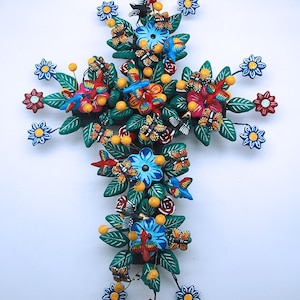 Può includere: Una croce decorativa artigianale con fiori, farfalle e bacche colorate. La croce presenta accenti rossi, blu e gialli, e foglie verdi. Il design è simmetrico e dettagliato, con un'estetica di arte popolare.