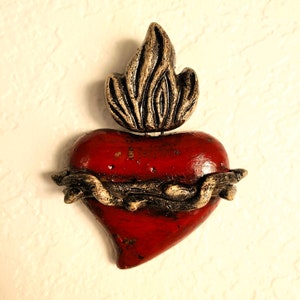 May include: A red heart with a gold flame on top and a thorny crown around it. The heart is made of clay and has a rustic, aged look.