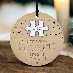 Puede incluir: Un adorno de madera con una pieza de rompecabezas blanca que dice "Papá" y el texto "Un pedazo de mi corazón vive en el cielo". El adorno está decorado con pequeñas estrellas.