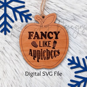 Puede incluir: Un adorno de madera con forma de manzana con el texto "Fancy Like Applebees" en una fuente marrón. El adorno tiene una cuerda para colgar.