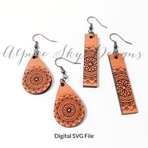 May include: Three pairs of wooden earrings with a mandala design. The earrings are in the shape of a teardrop and a rectangle. The earrings are a light brown color with a black mandala design.