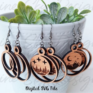 May include: Three pairs of wooden earrings with a circle design. The earrings feature a laser-cut design of a moon, stars, and mountains, a forest scene with stars, and a double circle design. The earrings are on a white surface with green plants in the background. Digital SVG File.