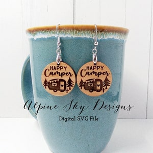May include: A pair of wooden earrings with a camper and trees design. The earrings are engraved with the text "Happy Camper".
