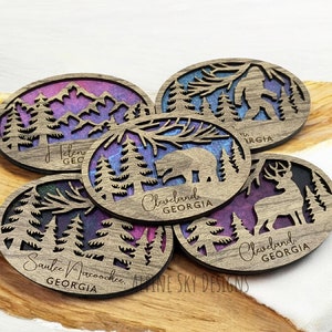 May include: Five wooden coasters with a nature theme. Each coaster features a different design, including a bear, a deer, a Bigfoot, and mountains. The coasters are painted with a purple and blue watercolor effect and have the text "Cleveland, Georgia" and "Sautee Nacoochee, Georgia" on them.