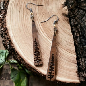 Puede incluir: Un par de pendientes de madera con un diseño de pino grabado con láser. Los pendientes son marrones y tienen una cuenta dorada en la parte superior del gancho.