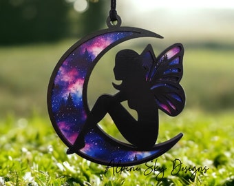 Puede incluir: Una silueta negra de un hada sentada en una luna creciente con estampado de galaxia. La luna cuelga de una cuerda.
