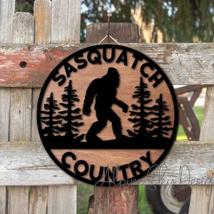 May include: A round wooden sign with the words "Sasquatch Country" in black lettering. A silhouette of a Sasquatch walking through a forest is in the center of the sign.