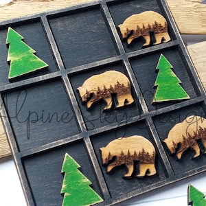 May include: A wooden tic-tac-toe game with a black background and green trees and brown bears with forest silhouettes. The game is made of wood and has a rustic look.