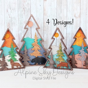 Può includere: Quattro decorazioni per l'albero di Natale in legno con diversi design. Ogni albero ha una scena diversa all'interno, tra cui un lupo, un orso, un cervo e un paesaggio montano. Gli alberi sono dipinti in tonalità di marrone, blu, verde e arancione. Il testo "4 Designs!" è scritto in nero su sfondo bianco sopra gli alberi.