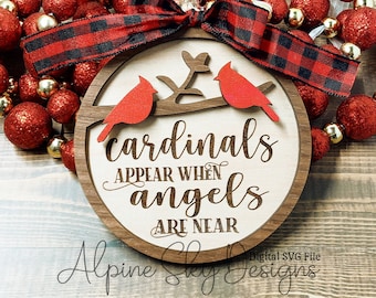 May include: A wooden Christmas ornament with a light wood center and a dark wood frame. The ornament features two red cardinal birds perched on a branch, with the words "cardinals appear when angels are near." A red and black plaid ribbon is attached.