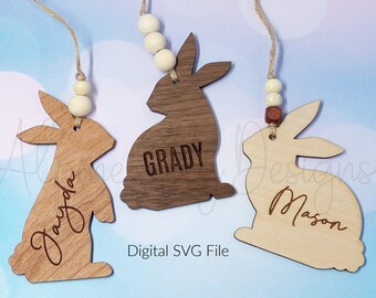 May include: Three wooden bunny ornaments with names engraved. Each bunny is a different shade of brown and tan. The bunnies are strung with twine and wooden beads. The text "Digital SVG File" is also visible.