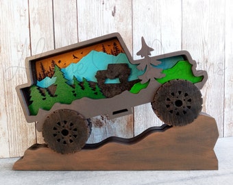 May include: A brown wooden cutout of a jeep driving up a hill with a mountain scene inside the jeep. The jeep has large tires and a green interior. The mountain scene is a blue and green landscape with trees and a brown sky.