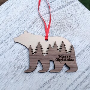 Puede incluir: Adorno navide&ntilde;o de madera con forma de oso. La parte superior es de color marr&oacute;n claro, la parte inferior es de color marr&oacute;n oscuro. Presenta un dise&ntilde;o de bosque recortado y la inscripci&oacute;n "Merry Christmas".
