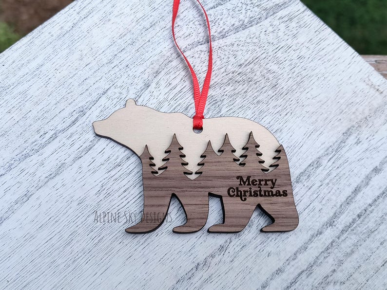 Puede incluir: Adorno navide&ntilde;o de madera con forma de oso. La parte superior es de color marr&oacute;n claro, la parte inferior es de color marr&oacute;n oscuro. Presenta un dise&ntilde;o de bosque recortado y la inscripci&oacute;n "Merry Christmas".