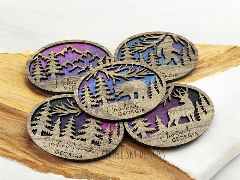 May include: Five wooden coasters with a nature theme. Each coaster features a different design, including a bear, a deer, a Bigfoot, and mountains. The coasters are painted with a purple and blue watercolor effect and have the text "Cleveland, Georgia" and "Sautee Nacoochee, Georgia" on them.