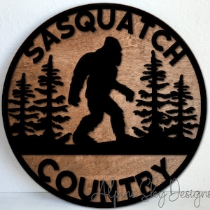 May include: A round wooden sign with a black silhouette of Bigfoot walking through a forest. The sign says "Sasquatch Country" in black lettering.