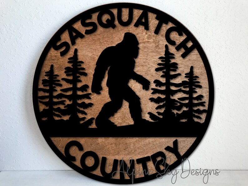 May include: A round wooden sign with a black silhouette of Bigfoot walking through a forest. The sign says "Sasquatch Country" in black lettering.