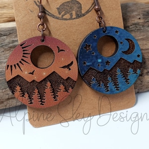 May include: A pair of wooden earrings with a mountain and sky design. One earring features a sunset with a sun and birds, while the other earring features a night sky with a crescent moon and stars.