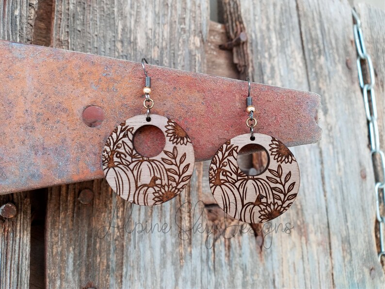 May include: A pair of wooden earrings with a pumpkin and sunflower design. The earrings are round and have a hole in the center. The design is laser-cut into the wood.