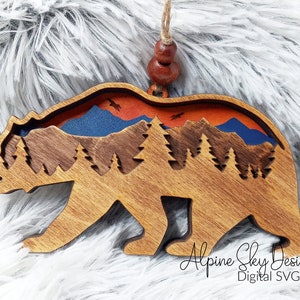Op de afbeelding: Houten berenversiering met een uitgesneden ontwerp van een bergketen, bomen en een zonsondergangshemel. De beer is bruin en de hemel is oranje, blauw en rood. De tekst "Alpine Sky Designs Digital SVG File" staat onderaan de versiering.