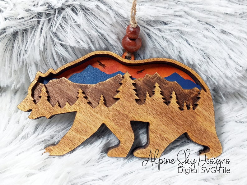 Op de afbeelding: Houten berenversiering met een uitgesneden ontwerp van een bergketen, bomen en een zonsondergangshemel. De beer is bruin en de hemel is oranje, blauw en rood. De tekst "Alpine Sky Designs Digital SVG File" staat onderaan de versiering.