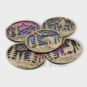 May include: Five wooden coasters with a nature theme. Each coaster features a different design with a bear, a Bigfoot, a deer, and two different forest scenes. The coasters are stained with a purple and blue gradient and have the text "Cleveland, Georgia" and "Sautee Nacoochee, Georgia" on them.