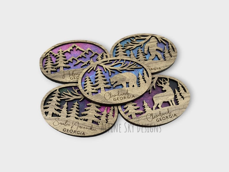 May include: Five wooden coasters with a nature theme. Each coaster features a different design with a bear, a Bigfoot, a deer, and two different forest scenes. The coasters are stained with a purple and blue gradient and have the text "Cleveland, Georgia" and "Sautee Nacoochee, Georgia" on them.