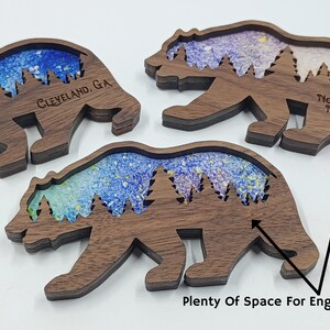 May include: Three wooden bear silhouette ornaments with a blue and purple watercolor background. The bears have a space for engraving on the bottom. The text "Cleveland, GA" is engraved on the left bear and "North Georgia Mountains" is engraved on the right bear.