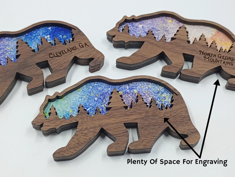 May include: Three wooden bear silhouette ornaments with a blue and purple watercolor background. The bears have a space for engraving on the bottom. The text "Cleveland, GA" is engraved on the left bear and "North Georgia Mountains" is engraved on the right bear.