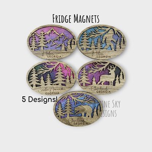 May include: Five wooden fridge magnets with a nature theme. Each magnet features a different design with a watercolor background and a silhouette of a mountain, a bear, a deer, a Bigfoot, and a forest. The magnets are oval shaped and have the name of a town in Georgia written on them.