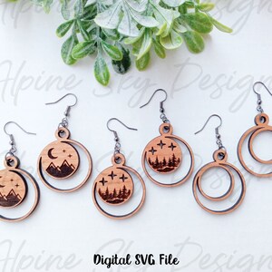 May include: Five pairs of wooden earrings with a moon and mountain design. The earrings are laser cut and have a natural wood finish. The design features a crescent moon with stars and a mountain range. Two of the earrings are simple circles.