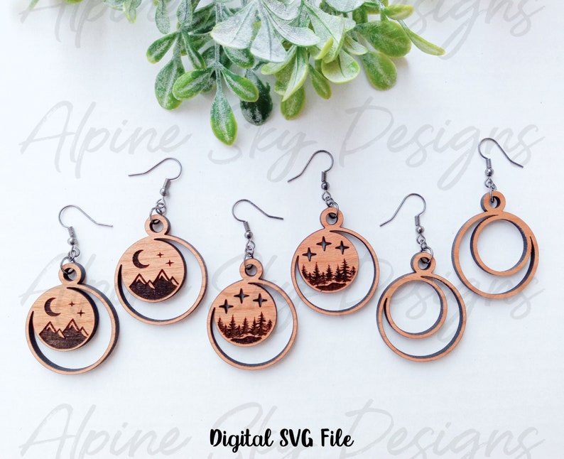 May include: Five pairs of wooden earrings with a moon and mountain design. The earrings are laser cut and have a natural wood finish. The design features a crescent moon with stars and a mountain range. Two of the earrings are simple circles.