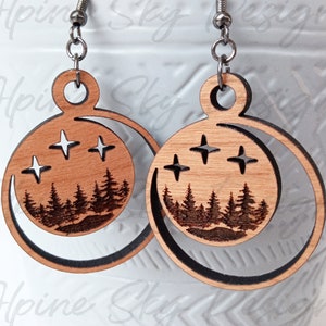 May include: A pair of wooden earrings with a crescent moon design. The earrings feature a silhouette of a forest with stars in the sky. The earrings are made of light brown wood and have silver hooks.