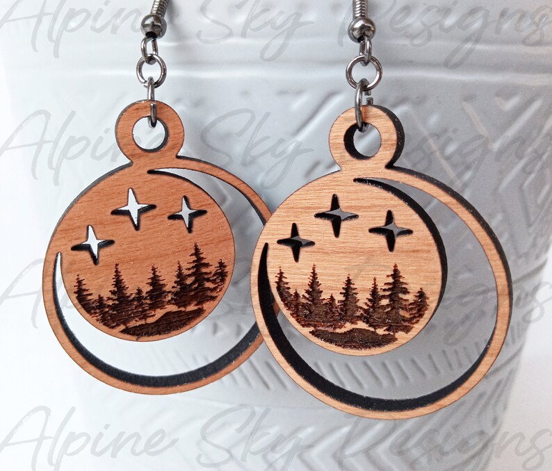 May include: A pair of wooden earrings with a crescent moon design. The earrings feature a silhouette of a forest with stars in the sky. The earrings are made of light brown wood and have silver hooks.