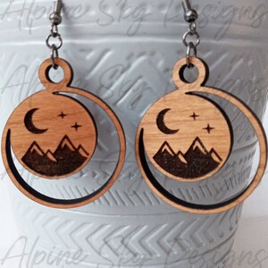 May include: A pair of wooden earrings with a crescent moon, stars, and mountain silhouette design. The earrings are made of light brown wood with a dark brown outline.