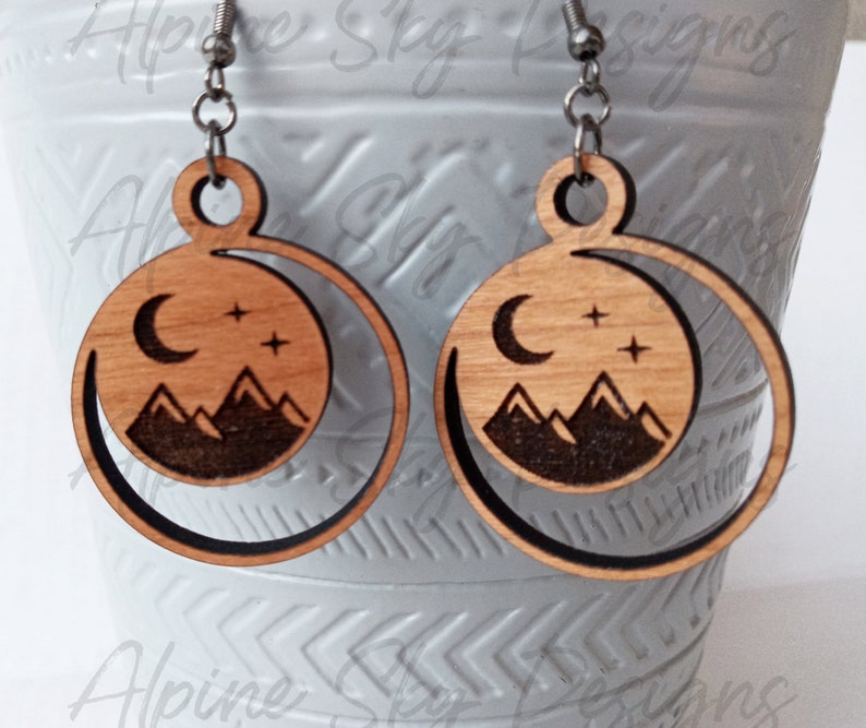 May include: A pair of wooden earrings with a crescent moon, stars, and mountain silhouette design. The earrings are made of light brown wood with a dark brown outline.