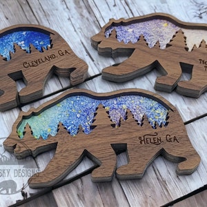 May include: Three wooden bear silhouette ornaments with a colorful resin inlay depicting a forest scene. The ornaments are engraved with the text "Cleveland, GA", "Helen, GA", and "North Georgia Mountains".