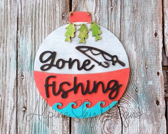 May include: A round wooden sign with a red, white, and blue design. The sign says "Gone Fishing" with a fishing rod and three fish. The sign is hanging on a wooden wall.