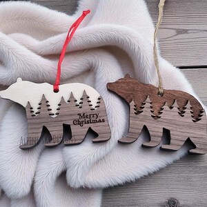 Puede incluir: Dos adornos de madera con forma de oso con un dise&ntilde;o de bosque. Un adorno tiene el texto "Merry Christmas". Los adornos son de color marr&oacute;n y crema, con cuerdas para colgar rojas y beige. Est&aacute;n sobre una superficie blanca y esponjosa.