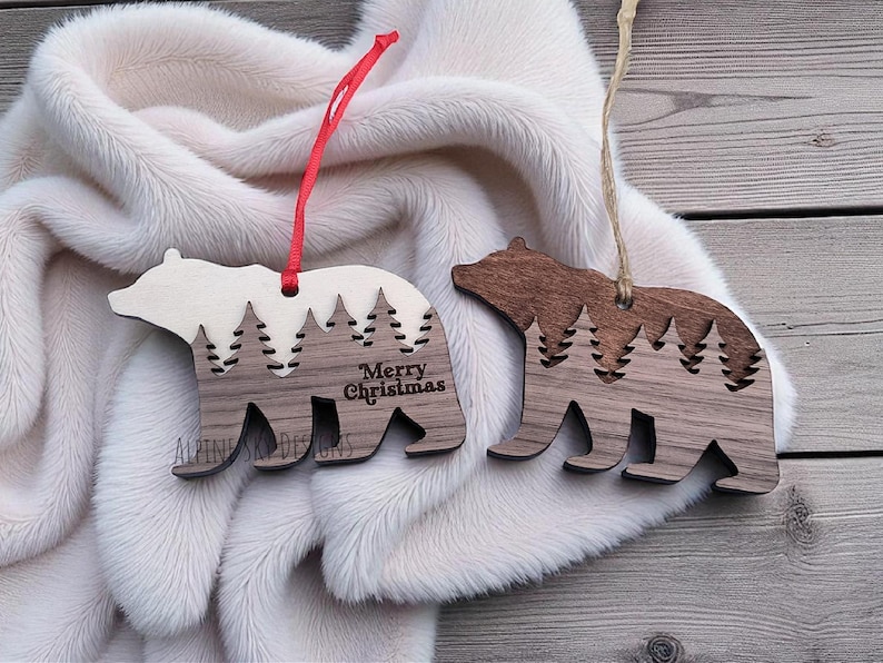 Puede incluir: Dos adornos de madera con forma de oso con un dise&ntilde;o de bosque. Un adorno tiene el texto "Merry Christmas". Los adornos son de color marr&oacute;n y crema, con cuerdas para colgar rojas y beige. Est&aacute;n sobre una superficie blanca y esponjosa.