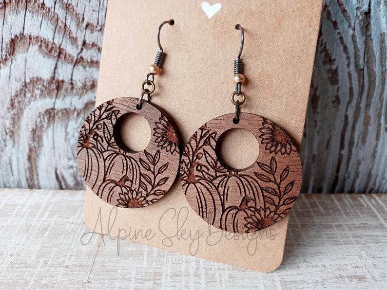 May include: A pair of wooden earrings with a round shape and a cutout in the center. The earrings are decorated with a laser-engraved design of pumpkins, sunflowers, and leaves. The earrings are brown in color and have a rustic look.