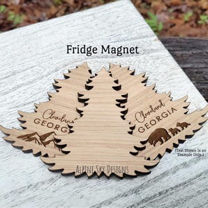 May include: Wooden fridge magnets in the shape of stylized trees. Each magnet features the text "Cleveland GEORGIA", a mountain range, and a bear family. The text "Fridge Magnet" and "Alpine Sky Designs" are also visible.