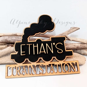 May include: A wooden train-shaped sign with the name "ETHAN'S" cut out in a light color, set against a black background. Below, the words "TRAIN STATION" are also cut out. The sign is placed on a white surface with driftwood.