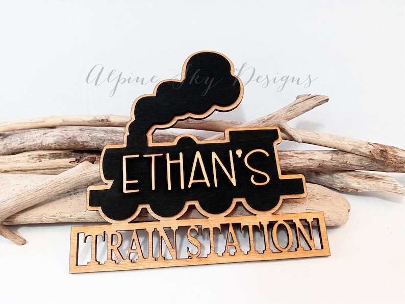 May include: A wooden train-shaped sign with the name "ETHAN'S" cut out in a light color, set against a black background. Below, the words "TRAIN STATION" are also cut out. The sign is placed on a white surface with driftwood.
