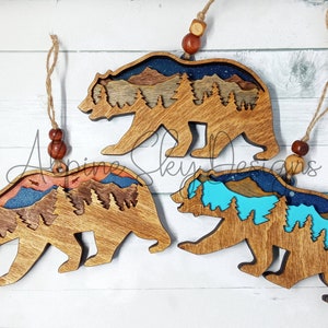 Op de afbeelding: Drie houten berenversieringen met een berg- en boslandschap erin. De beren zijn bruin en de landschappen zijn geschilderd in blauw, turquoise, oranje en bruin.