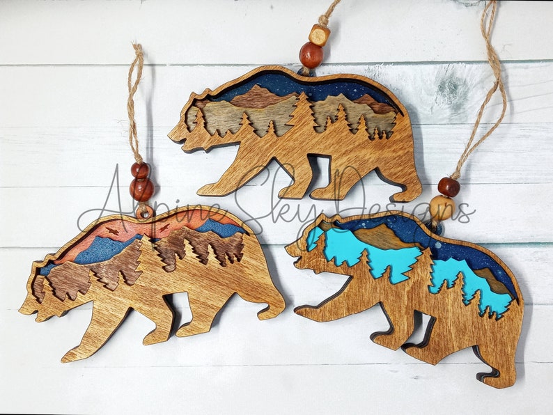 Op de afbeelding: Drie houten berenversieringen met een berg- en boslandschap erin. De beren zijn bruin en de landschappen zijn geschilderd in blauw, turquoise, oranje en bruin.