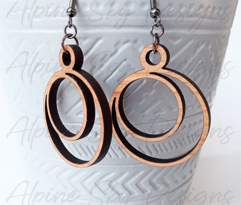 May include: A pair of wooden earrings with a double circle design. The earrings are made of natural wood with a dark brown stain. The earrings are attached to silver hooks.