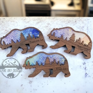 May include: Three wooden bear silhouette magnets with a colorful glitter background. The magnets feature the text "Cleveland, GA", "Helen, GA", and "North Georgia Mountains".