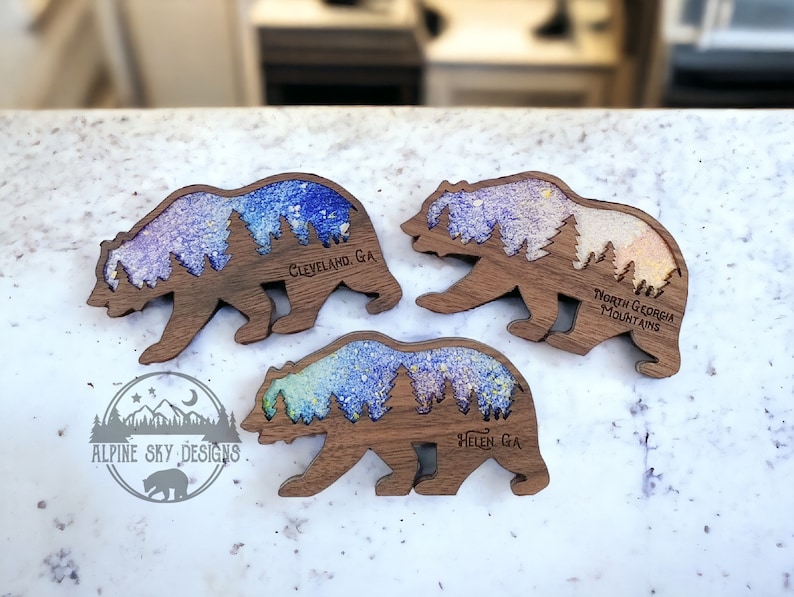 May include: Three wooden bear silhouette magnets with a colorful glitter background. The magnets feature the text "Cleveland, GA", "Helen, GA", and "North Georgia Mountains".