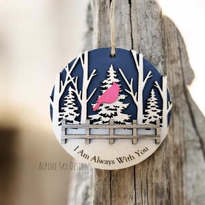 Puede incluir: Un adorno navide&ntilde;o de madera con fondo azul y una silueta blanca de una escena de bosque. Un p&aacute;jaro cardenal rosa est&aacute; posado en una rama. El adorno dice "I Am Always With You" y "Alpine Sky Designs".